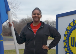 Photo of a UAW Local 163 member holding a UAW flag, standing in front of the local union hall. Local 163 members work at Detroit Axle.