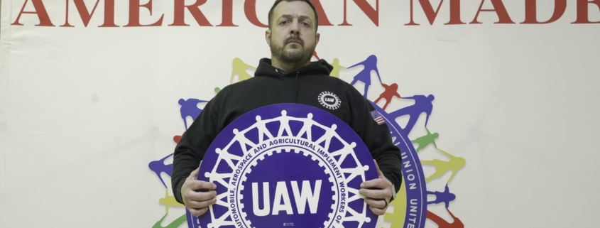 Photo of a Local 933 member holding a UAW sign with a "American Made, Union Proud" back drop.