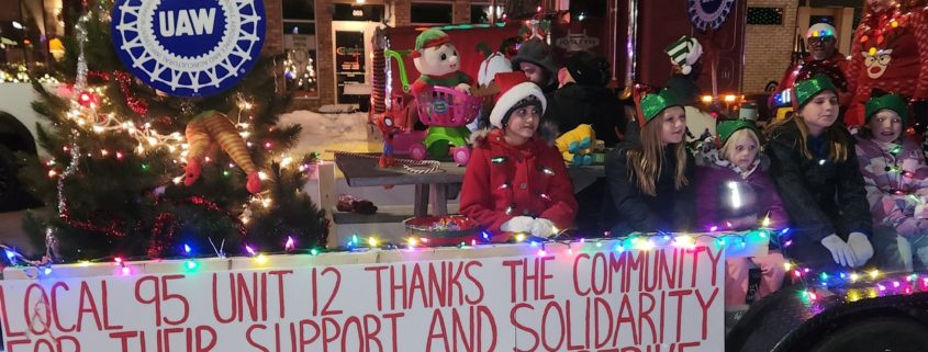 UAW wheel sign, thank you for support and solidarity banner, Christmas lights, photo of children and adults posing in front of neighborhood homes in winter attire.