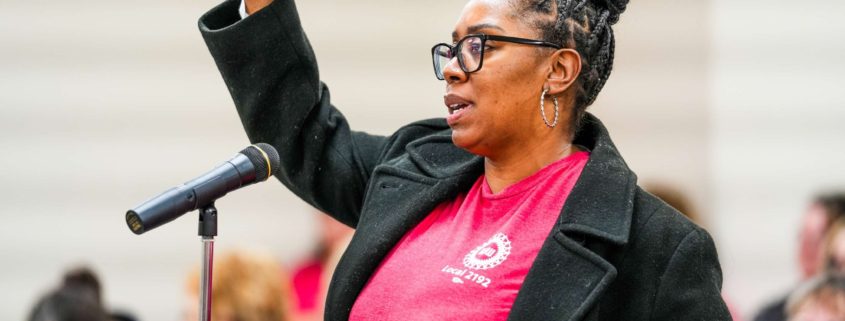 Member standing at mic wearing a red UAW t-shirt and black blazer, inside a UAW local union hall.